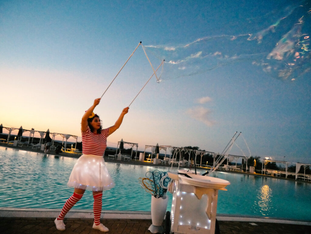 LELLA BRETELLA è un clown giocherellone che adora realizzare sculture di palloncini da donare ai bambini. In questo modo intrattiene piccoli e grandi durante l’accoglienza di un evento. Da notare l’originalità del personaggio che si presta benissimo sia per eventi in piazza che privati. Si presenta già con il vestito pieno di figure di palloncini e continua a farne insieme agli ospiti intrattenendo con gags gioiose.